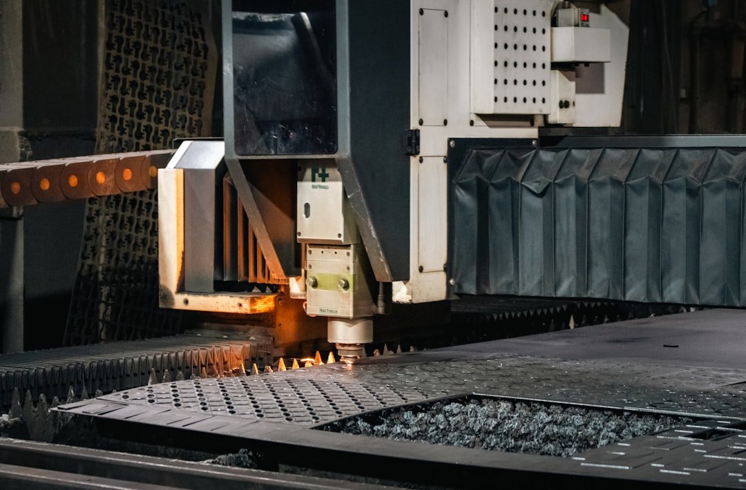 A close-up captures the action of an industrial CNC laser/plasma machine actively cutting a metal sheet. The cutting head is visible, emitting a small spark or glow of heat as it moves across the metal surface supported by a spiked cutting bed.